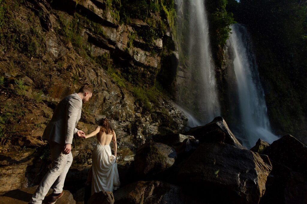 Couple climbing wet rocks toward twin waterfalls in hard sunlight