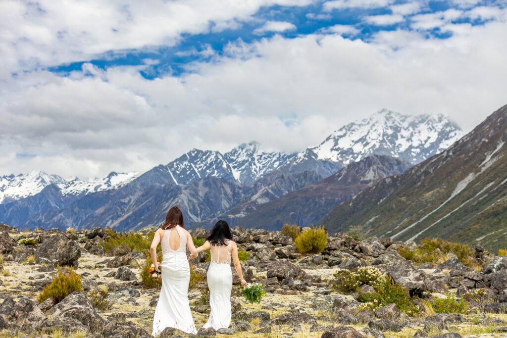 Two brides in open-back lace dresses walking toward snow-covered peaks