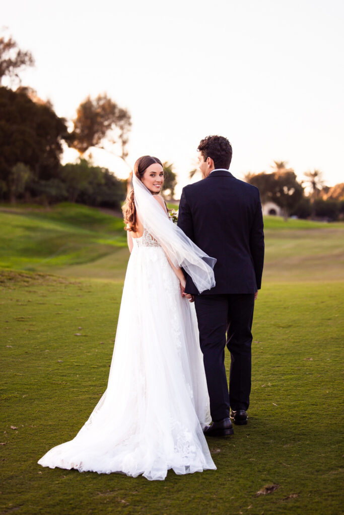 Bride looking back over her shoulder at sunset on a golf course as her veil lifts