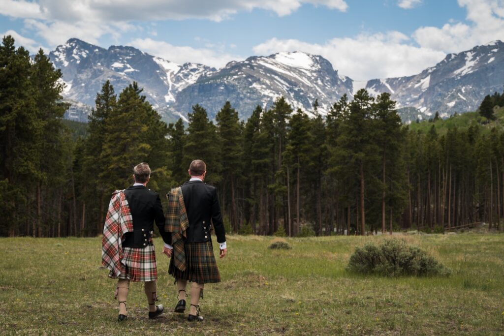 Two grooms in kilts walking hand in hand across a mountain meadow