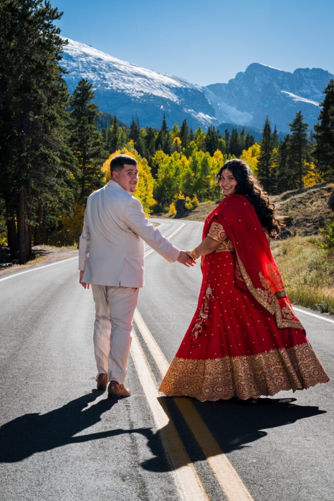 Couple on a mountain road among yellow aspens, both looking back, bride in a red lehenga