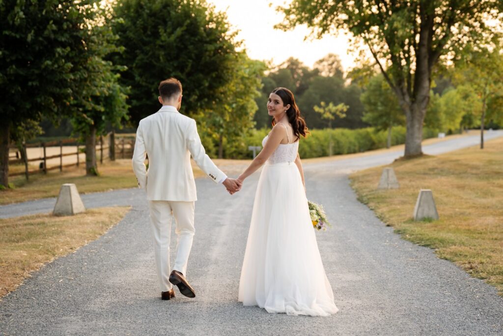 Couple walking away down a tree-lined gravel drive as the bride looks back