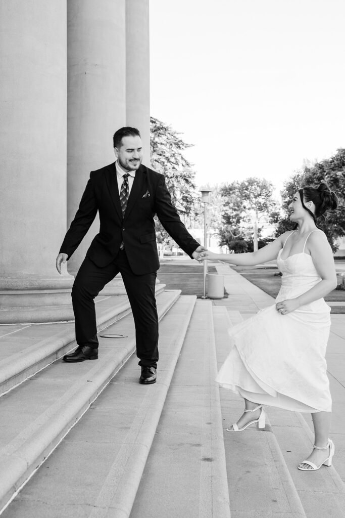 Black and white photo of a groom leading the bride up stone steps as she swings her skirt