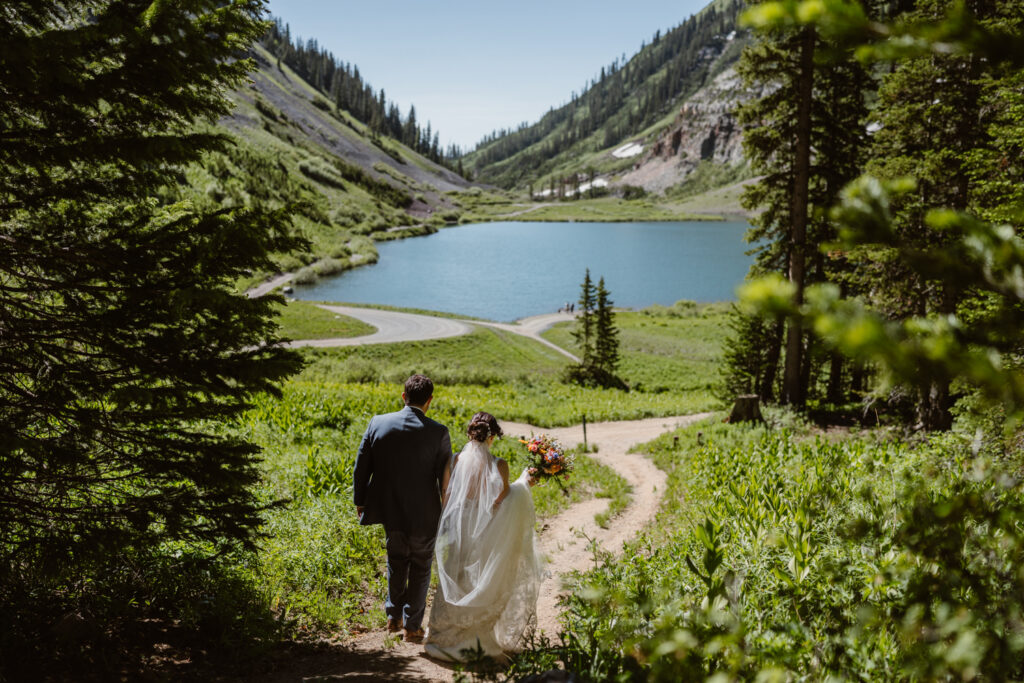 Couple walking down a trail toward an alpine lake framed by evergreen branches